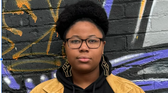 Photo of Allison Rolle - African diasporic person with glasses wearing a black hoodie standing in front of a black brick wall.