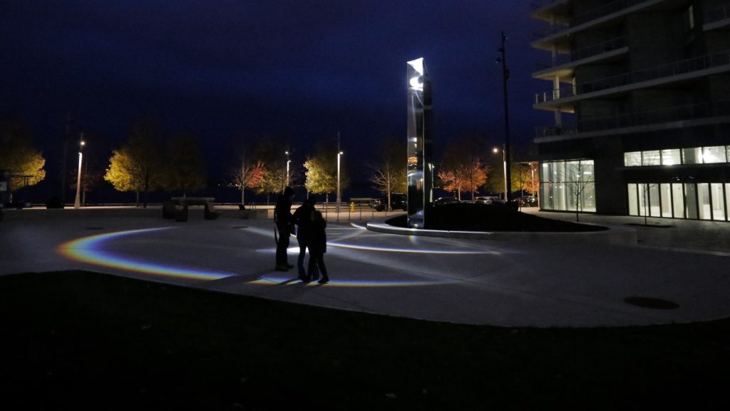 A tall silver cylinder structure that emits rainbows onto the ground